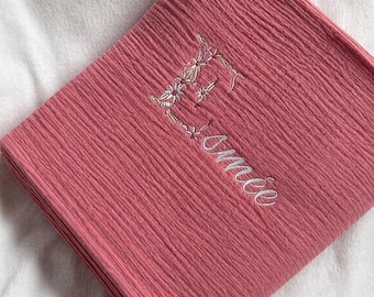 May include: A folded pink muslin blanket with a textured, crinkled surface. The fabric features white embroidered floral detailing forming the letter E, followed by the white embroidered cursive text Aimée positioned below the letter.