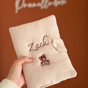 Peut inclure: Une housse en tissu mousseline beige avec le prénom Zack brodé en fil marron foncé au-dessus d'un petit motif d'ours en peluche marron foncé. La housse rectangulaire comprend une patte latérale avec un bouton-pression blanc de fermeture.
