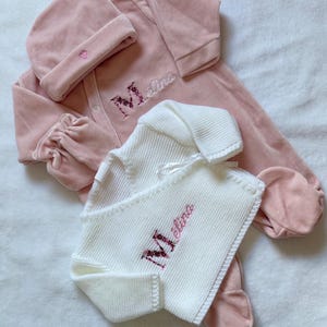 Peut inclure: Ensemble de vêtements pour bébé rose clair. L'ensemble comprend une combinaison à pieds, un bonnet assorti avec un détail cœur et une couverture en tricot blanc. Le nom "Molina" est brodé en rose sur la combinaison et la couverture.