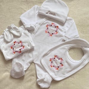 Peut inclure: Ensemble de vêtements de bébé blanc comprenant une combinaison à pieds, un bavoir, un chapeau et des moufles. Chaque article est brodé du nom "Lexie" et d'une couronne florale rose et orange. L'ensemble est en matière douce.