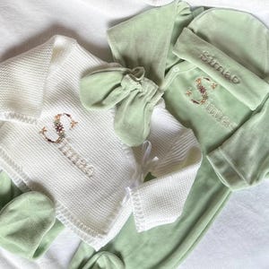 Peut inclure: Un ensemble de vêtements pour bébé en vert clair et blanc. L'ensemble comprend une couverture en tricot, un body, un chapeau et des moufles. Le nom "Simao" est brodé avec un motif floral sur la couverture et le body.