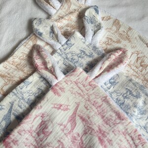 May include: Three baby blankets with bunny ear designs. The blankets feature a toile pattern with giraffes and airplanes in shades of pink, blue, and tan. The ears are white and fluffy, adding a soft texture to the design.