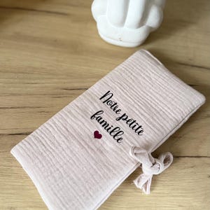 May include: A light pink, rectangular fabric item with the text "Notre petite famille" and a small heart. The fabric is textured and tied with a knot. A white, decorative, abstract sculpture is in the background.