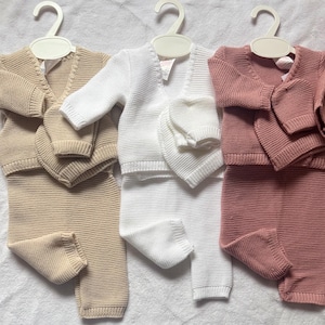 Può includere: Tre set di abbigliamento in maglia, ciascuno composto da un cardigan a maniche lunghe con scollo a V e pantaloni abbinati. I set sono nei colori beige, bianco e rosa antico. Ogni capo presenta una consistenza uniforme a maglia a coste.