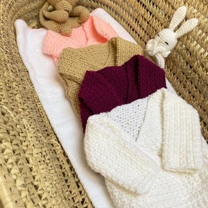 Op de afbeelding: Vier gehaakte babytruien in een rieten mand. De truien zijn in de kleuren roze, beige, bordeauxrood en wit.