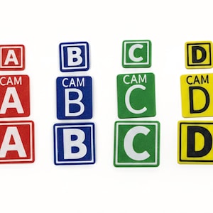Könnte beinhalten: Quadratische Aufkleber mit den Buchstaben A, B, C und D in Rot, Blau, Grün und Gelb. Einige Aufkleber tragen das Wort "CAM" über dem Buchstaben. Ideal für Bildungs- oder Organisationszwecke.