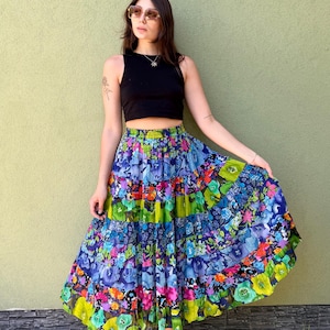 Op de afbeelding: Een zwarte mouwloze crop top gecombineerd met een lange, gelaagde, veelkleurige rok met een levendig bloemenpatroon in tinten blauw, groen, roze en geel. De rok heeft een geplooide elastische tailleband en een vol, zwierig silhouet.
