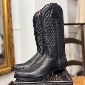 Op de afbeelding: Een paar zwarte leren cowboylaarzen met ingewikkelde stikselontwerpen. De laarzen hebben spitse tenen en gestapelde hakken. Ze worden gepresenteerd op een decoratieve metalen standaard, wat hun klassieke Western-stijl benadrukt.
