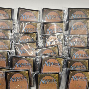 May include: A collection of Magic: The Gathering trading cards, each enclosed in a clear plastic protective sleeve. The cards are displayed with their backs facing forward, showing the iconic tan and blue design with the Magic logo and Deckmaster text.