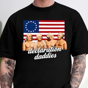 May include: A black short-sleeved t-shirt featuring a graphic of four shirtless historical figures standing in front of an American flag with thirteen stars. Below the figures, the white text reads "declaration daddies" in a cursive script font.