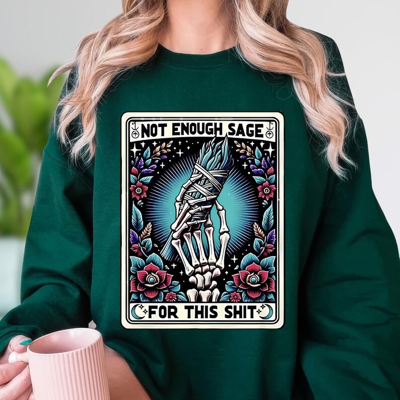 Puede incluir: Sudadera verde oscuro con un gr&aacute;fico que presenta una mano esquel&eacute;tica sosteniendo un manojo de salvia. El dise&ntilde;o incluye el texto "NOT ENOUGH SAGE" y "FOR THIS SHIT" en un marco decorativo con elementos florales y celestiales.