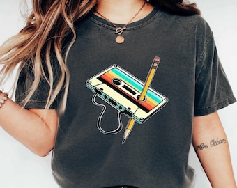 May include: Dark grey t-shirt featuring a retro cassette tape design with a pencil piercing through it. The cassette has a colorful striped design. The shirt is a casual wear item.