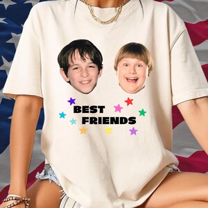 May include: Cream-colored t-shirt featuring two smiling faces and the words "BEST FRIENDS" in black. Colorful star graphics surround the text. The shirt is a casual style.