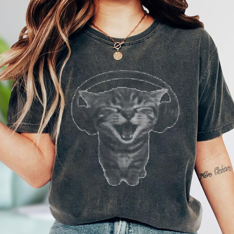 May include: Dark gray t-shirt featuring a pixelated cat wearing headphones. The cat graphic is in white, creating a contrast against the shirt's color. The shirt has a vintage, worn look.