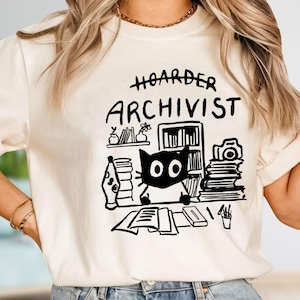 May include: A cream-colored short-sleeve t-shirt featuring a black line-art graphic of a cat sitting at a desk with books, a camera, and a lava lamp. The text above the illustration reads HOARDER ARCHIVIST in a hand-drawn, bold black font style.