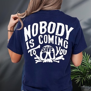 a person wearing a navy blue t-shirt with the text "NOBODY IS COMING TO SAVE YOU" printed on it.