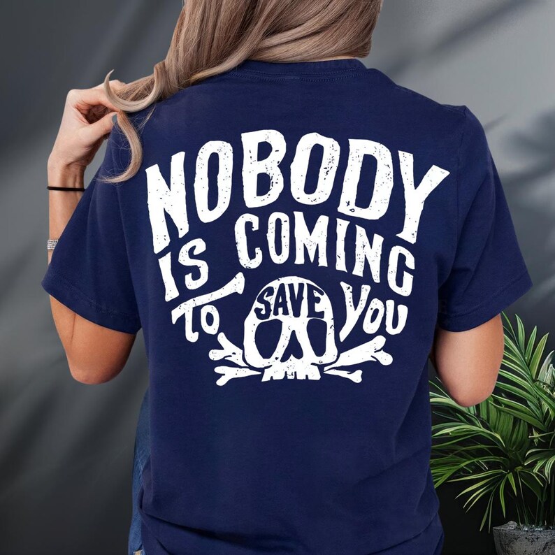 a person wearing a navy blue t-shirt with the text "NOBODY IS COMING TO SAVE YOU" printed on it.