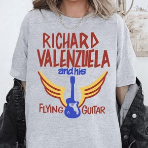 May include: Gray t-shirt with red and yellow graphic design. The graphic features a guitar with wings and the text "Richard Valenzuela and his Flying Guitar".