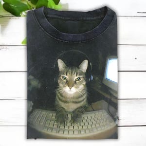 May include: Black t-shirt featuring a tabby cat wearing headphones and using a keyboard. The cat is in front of a computer monitor. The shirt has a crew neck and is made of a soft material. The image is a humorous take on a gamer cat.