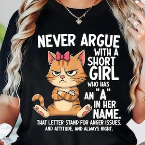 May include: Black t-shirt featuring a cartoon cat with an angry expression and a pink bow. The text reads "NEVER ARGUE WITH A SHORT GIRL WHO HAS AN 'A' IN HER NAME." The shirt's message continues with "THAT LETTER STAND FOR ANGER ISSUES, AND ATTITUDE, AND ALWAYS RIGHT."
