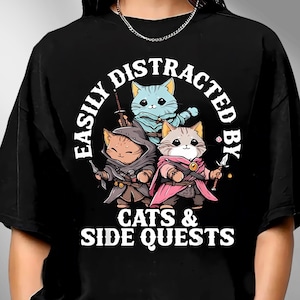 May include: Black t-shirt with a graphic featuring three cartoon cats dressed as fantasy characters. The text reads "EASILY DISTRACTED BY CATS & SIDE QUESTS" in a circular design.