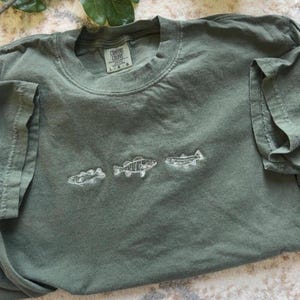 May include: An olive green t-shirt with a crew neck and short sleeves. The shirt has three embroidered white fish across the front. The label reads "Comfort Colors".