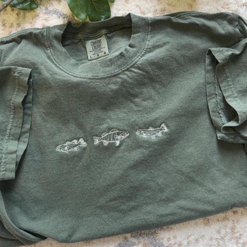 May include: An olive green t-shirt with a crew neck and short sleeves. The shirt has three embroidered white fish across the front. The label reads "Comfort Colors".