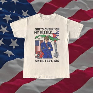 May include: Off-white t-shirt with a graphic featuring a man in a suit, a missile, and the text "SHE'S CUBIN' ON MY MISSILE UNTIL I CRY, SIS." The design includes the American and Cuban flags.