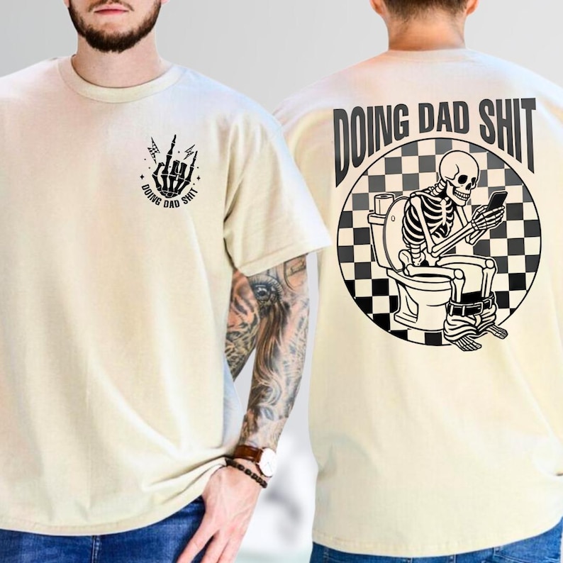 May include: Beige t-shirt with black graphic designs. The front features a skeleton hand with the text "DOING DAD SHIT". The back displays a skeleton on a toilet with a phone, surrounded by text and a checkered pattern.