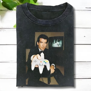 May include: Dark gray t-shirt featuring a graphic of a man in a tuxedo holding a gray video game controller. The image on the shirt includes a television screen. The shirt is made of a soft material.