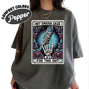 Puede incluir: Camiseta Comfort Colors color pimienta. La camiseta presenta un gr&aacute;fico con el texto "NOT ENOUGH SAGE FOR THIS SHIT" y una mano esquel&eacute;tica sosteniendo salvia. El gr&aacute;fico est&aacute; rodeado de elementos florales.