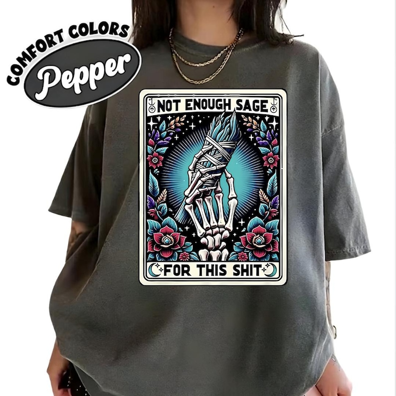 Puede incluir: Camiseta Comfort Colors color pimienta. La camiseta presenta un gr&aacute;fico con el texto "NOT ENOUGH SAGE FOR THIS SHIT" y una mano esquel&eacute;tica sosteniendo salvia. El gr&aacute;fico est&aacute; rodeado de elementos florales.