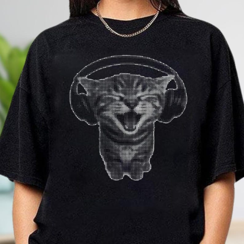May include: Black t-shirt featuring a graphic of a cat wearing headphones. The cat's face is rendered in a pixelated style, with its mouth open in a wide grin. The headphones are also depicted in a pixelated design, creating a unique visual effect.