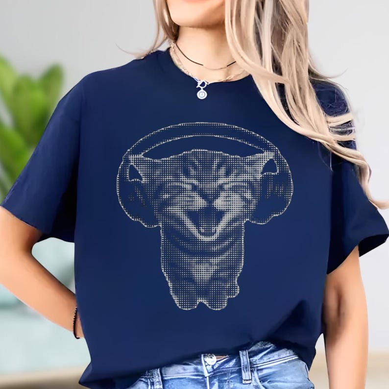 May include: Navy blue t-shirt featuring a white graphic of a cat wearing headphones. The cat's face is smiling, and the design is made up of small dots. The shirt has short sleeves and a crew neck.