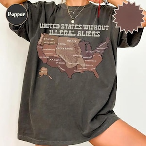 May include: Dark gray t-shirt featuring a map of the United States with the text "UNITED STATES WITHOUT ILLEGAL ALIENS." The map highlights various Native American tribes. The shirt is a casual wear item.