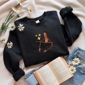May include: Black sweatshirt with an embroidered design of a seated figure with outstretched arms and three yellow stars. The sweatshirt is paired with blue jeans, a book, and daisies.