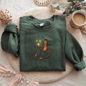 May include: Green sweatshirt with an embroidered cartoon figure and three yellow stars. The figure is brown with long limbs and a relaxed pose. The sweatshirt is folded on a woven mat, with a coffee cup and decorative items in the background.