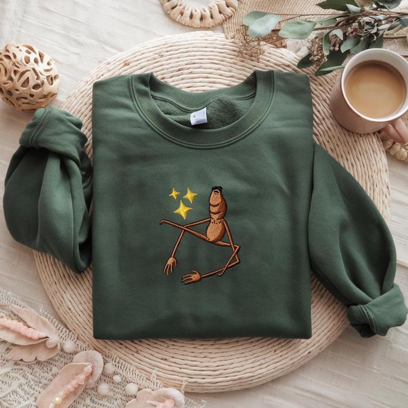 May include: Green sweatshirt with an embroidered cartoon figure and three yellow stars. The figure is brown with long limbs and a relaxed pose. The sweatshirt is folded on a woven mat, with a coffee cup and decorative items in the background.