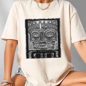 May include: Cream-colored t-shirt featuring a detailed black and white graphic of a stylized face within a rectangular frame. The face has intricate patterns and designs, creating a textured appearance. The shirt is a casual style, suitable for everyday wear.
