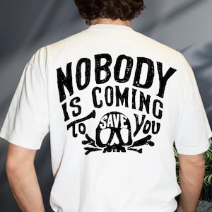 the back of a person wearing a white t-shirt with a black graphic design featuring the text "NOBODY IS COMING SAVE YOU".