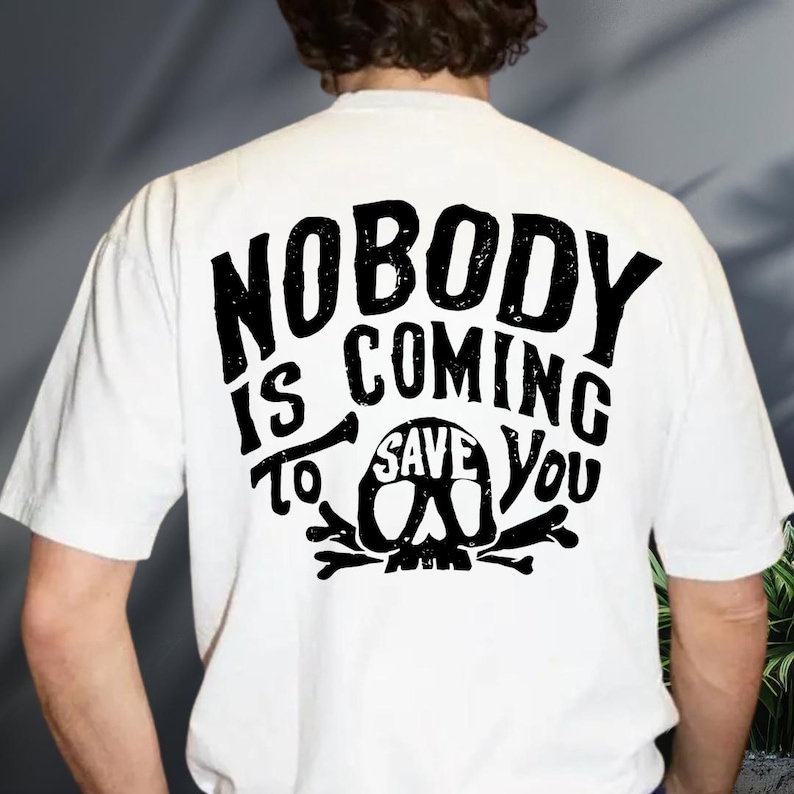the back of a person wearing a white t-shirt with a black graphic design featuring the text "NOBODY IS COMING SAVE YOU".