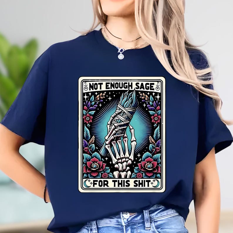 Puede incluir: Camiseta azul marino con un dise&ntilde;o que incluye el texto "NOT ENOUGH SAGE FOR THIS SHIT". El dise&ntilde;o muestra una mano esquel&eacute;tica sosteniendo un manojo de salvia, rodeada de flores y elementos celestiales.