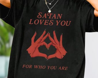 May include: Black t-shirt with the text "SATAN LOVES YOU" in red, above a graphic of red hands forming a heart shape. Below the graphic, the text reads "FOR WHO YOU ARE".