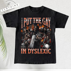 May include: Black t-shirt with a graphic design featuring skeleton figures, flames, and the text "I PUT THE GAY IN DYSLEXIC." The design includes a gun with the word "BAD ASS" on it.