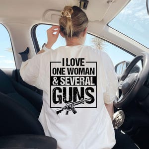 May include: White t-shirt with a black graphic on the back. The graphic includes the text "I LOVE ONE WOMAN & SEVERAL GUNS" inside a rectangular frame, with an illustration of a rifle below the text. The person is sitting in a car.