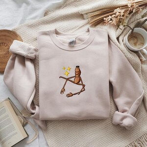 May include: A light pink sweatshirt with a crew neck and long sleeves. The sweatshirt features an embroidered cartoon character with brown limbs and a brown body, with three yellow stars above it. The sweatshirt is on a beige blanket.