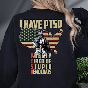 May include: Black sweatshirt with the text "I HAVE PTSD" above a graphic of the American flag and a soldier. Below the soldier, the text reads "PRETTY TIRED OF STUPID DEMOCRATS" in a distressed font.