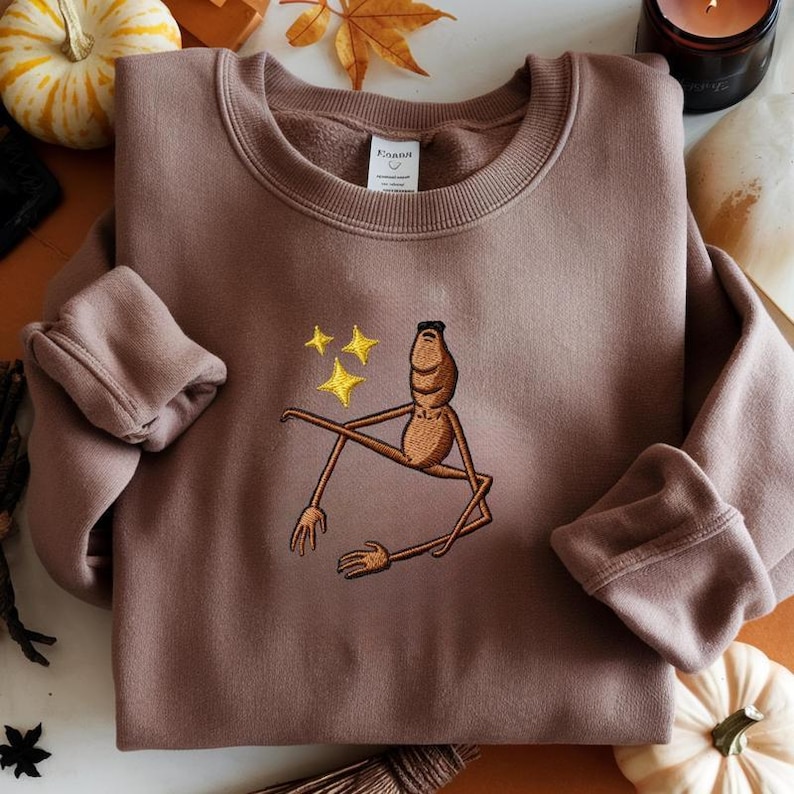 May include: A mauve-colored sweatshirt with an embroidered design of a whimsical, anthropomorphic creature with outstretched limbs and three yellow stars. The sweatshirt has a crew neck and long sleeves.