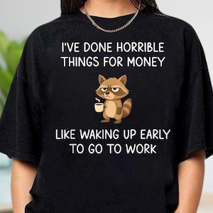 May include: Black t-shirt with the text "I'VE DONE HORRIBLE THINGS FOR MONEY" and "LIKE WAKING UP EARLY TO GO TO WORK". A cartoon raccoon holding a coffee cup is in the center.