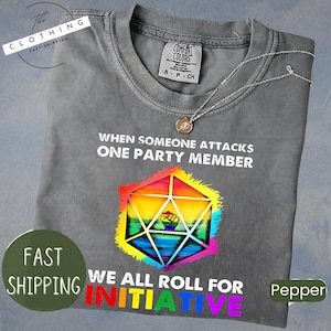 May include: A gray t-shirt with the text "WHEN SOMEONE ATTACKS ONE PARTY MEMBER WE ALL ROLL FOR INITIATIVE". The shirt features a colorful geometric design with a fist inside. A silver necklace is draped across the shirt.